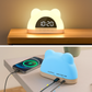Un Réveil Digital pour Enfant Coloré de Doux Reveils en forme d'ours brille doucement à 10h20 sur une table de nuit, chargeant un smartphone via des câbles arrière, avec une lumière bleue confortable et une tasse en arrière-plan.