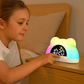 Une jeune fille en chemise blanche est assise sur un lit et touche le Doux Reveils Réveil Digital pour Enfant Coloré avec des lumières en forme d'ours sur une table de nuit en bois.
