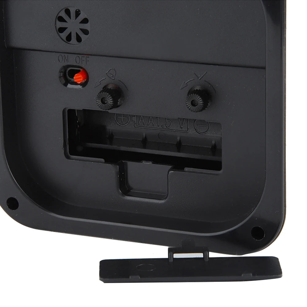 Vue rapprochée de l’arrière d’un réveil carré noir avec compartiment à piles et interrupteur rouge ON/OFF.