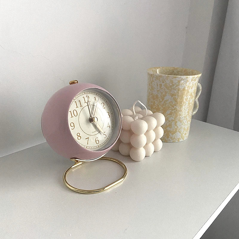 Réveil Vintage Silencieux Rond rose pastel sur support doré, posé près d’une bougie et d’une tasse jaune.